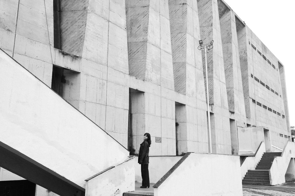 A woman stands outside a brutalist concrete building, looking upward.