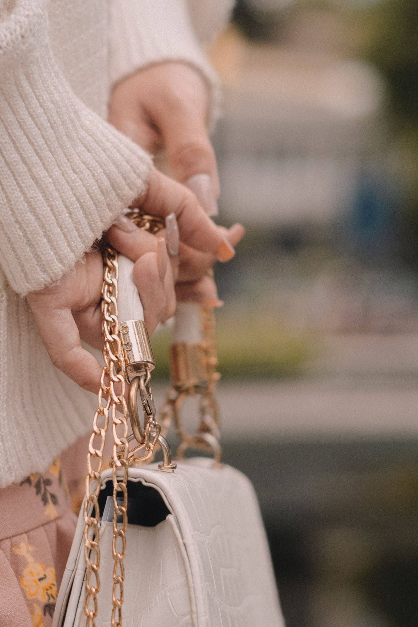 Close view of hands in pale knit sleeves holding the gold-colored chain straps of a structured white textured handbag.