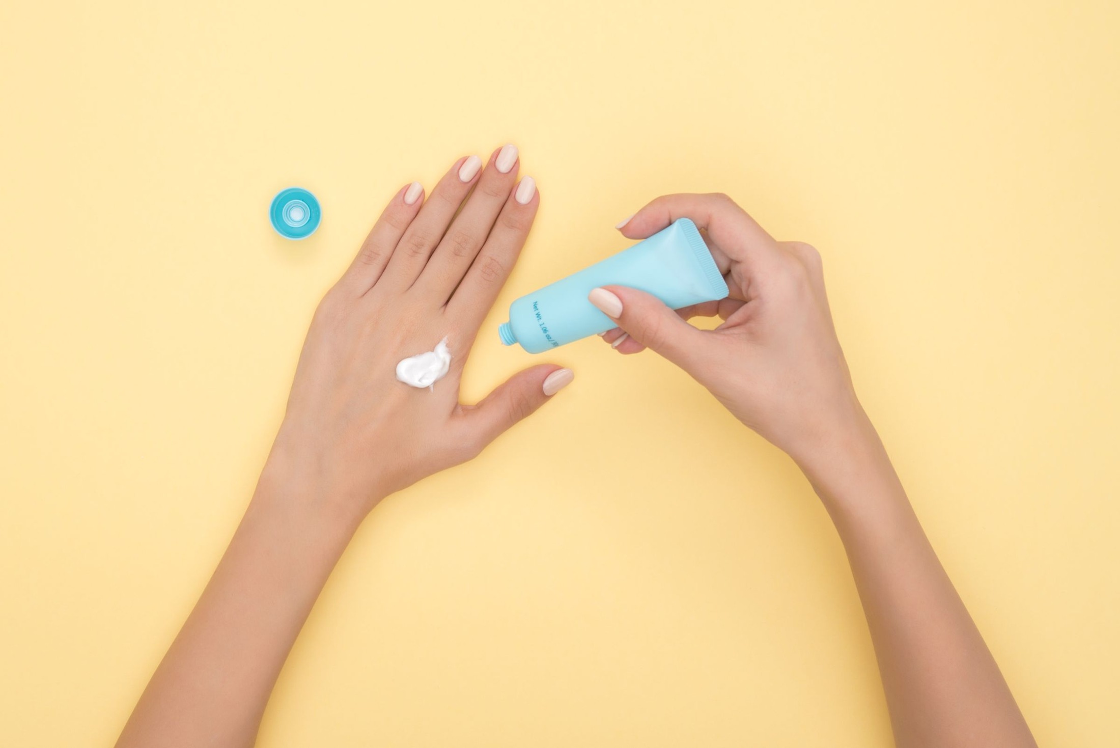 Hands dispensing white hand cream from a light-blue tube onto the back of a hand on a yellow surface, with the blue cap nearby.