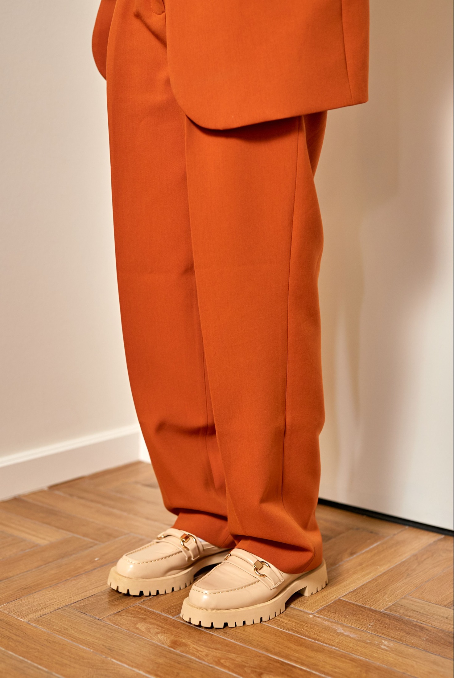 Orange tailored trouser hems rest over cream chunky-soled loafers on a wooden floor, showing front folds and the relationship between long hems and raised soles.