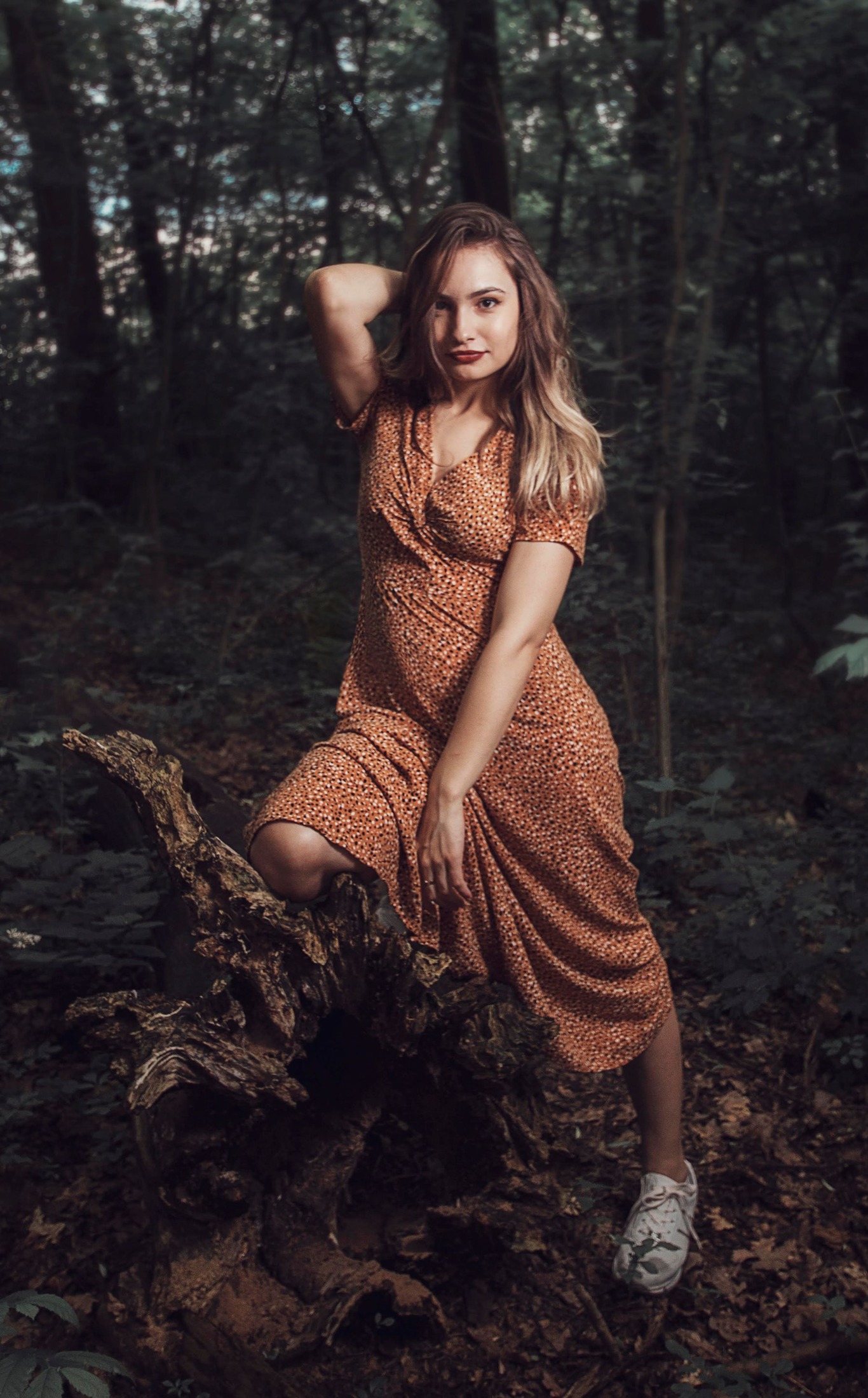 Woman in a rust-colored small-print midi dress and light sneakers, posing with one knee bent in woodland.
