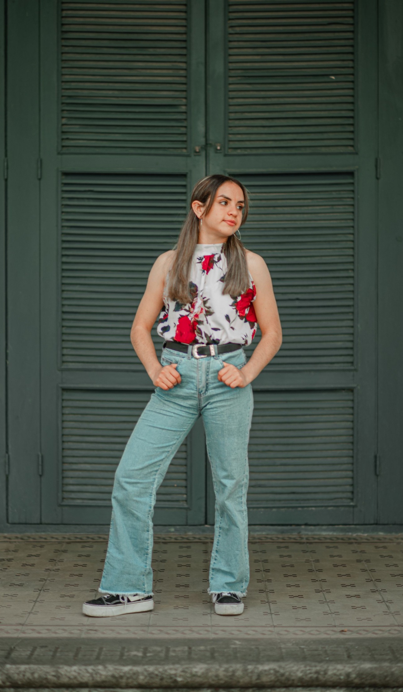 Woman wearing a sleeveless white blouse with red floral print, light-wash jeans, black belt and canvas sneakers before green shutters.