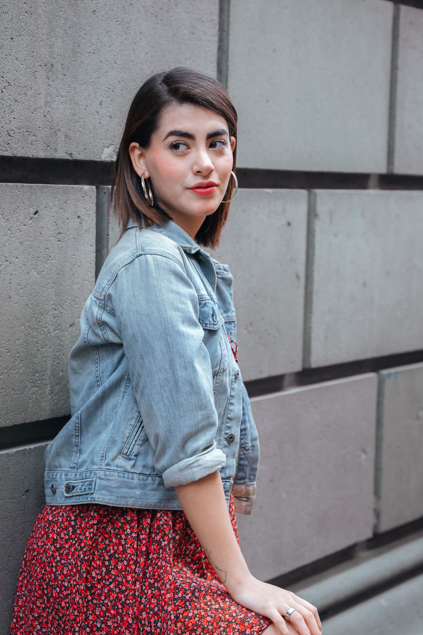 Woman wearing a cropped light-blue denim jacket with rolled sleeves over a small red floral-print dress beside a gray block wall.