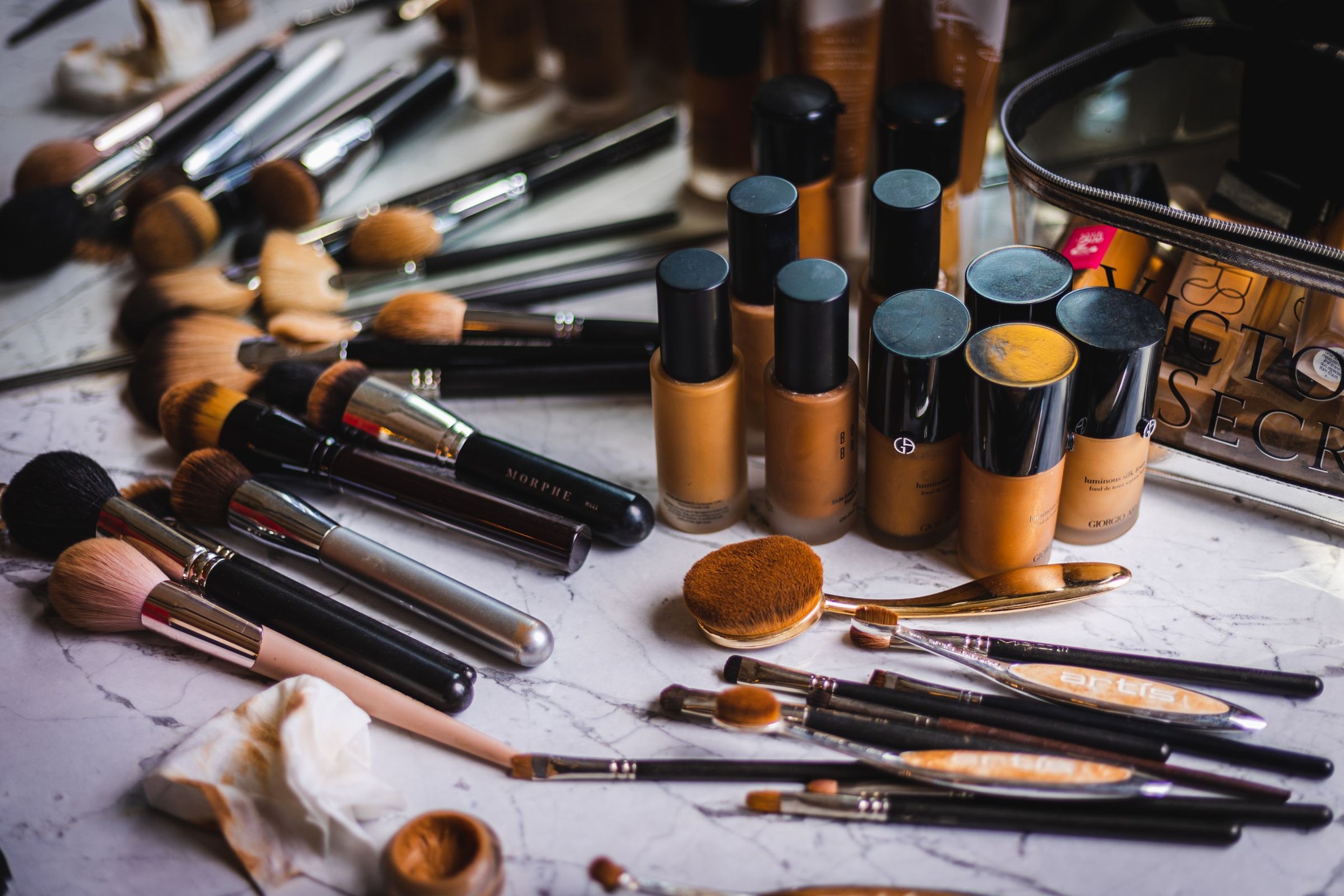 Several liquid foundation bottles in visibly different beige and brown shades standing among makeup brushes on a white work surface.