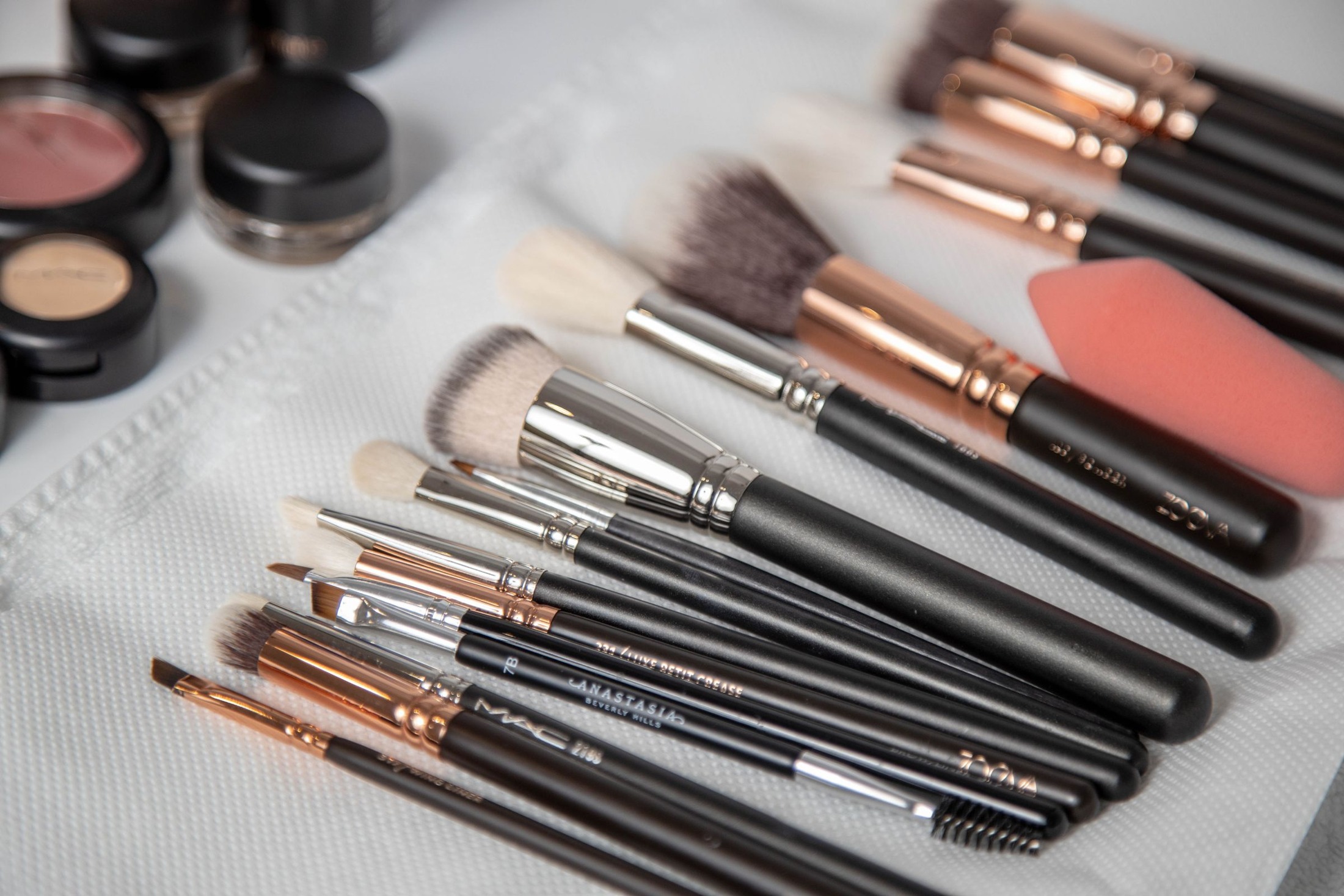 Makeup brushes lying flat on white toweling beside cosmetic pots and a peach sponge; illustrates a flat tool layout, not active washing or verified freshly washed brushes.