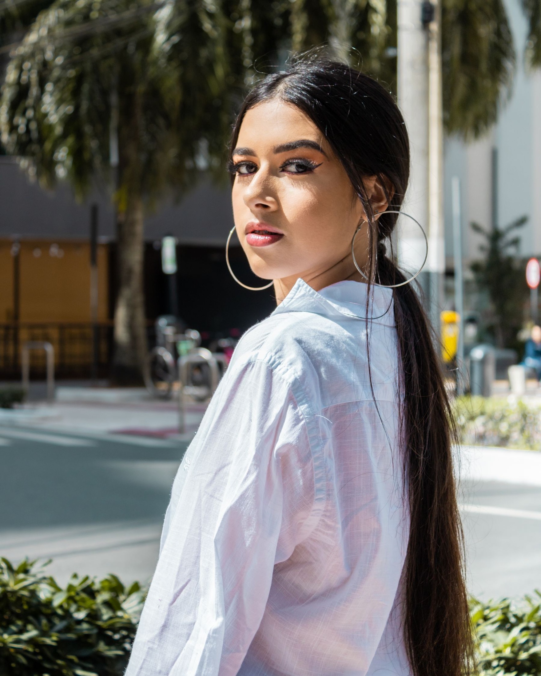 Woman with dark tied-back hair wearing large thin hoop earrings and a white collared shirt outdoors; the collar and hoop length are visible together.