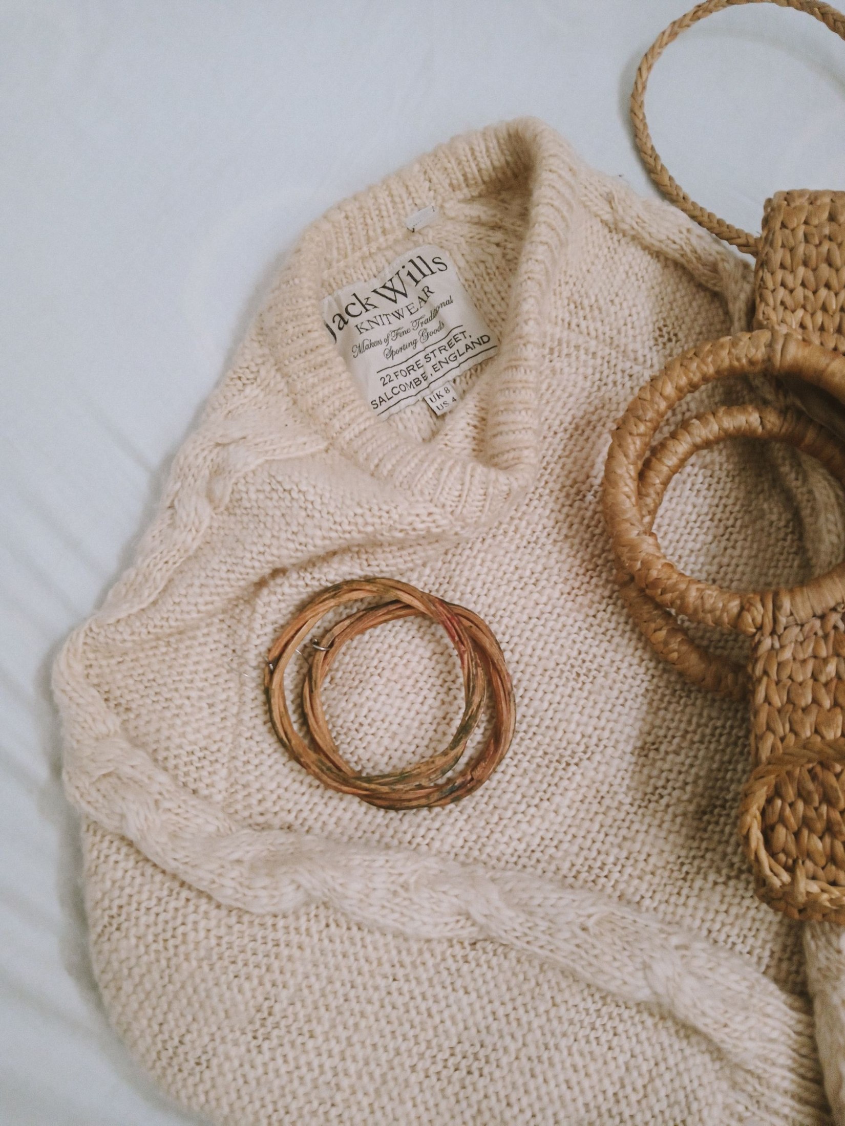 Cream knit sweater laid flat with its neck brand label visible beside woven accessories; illustrates knit construction and label location, not readable care instructions.