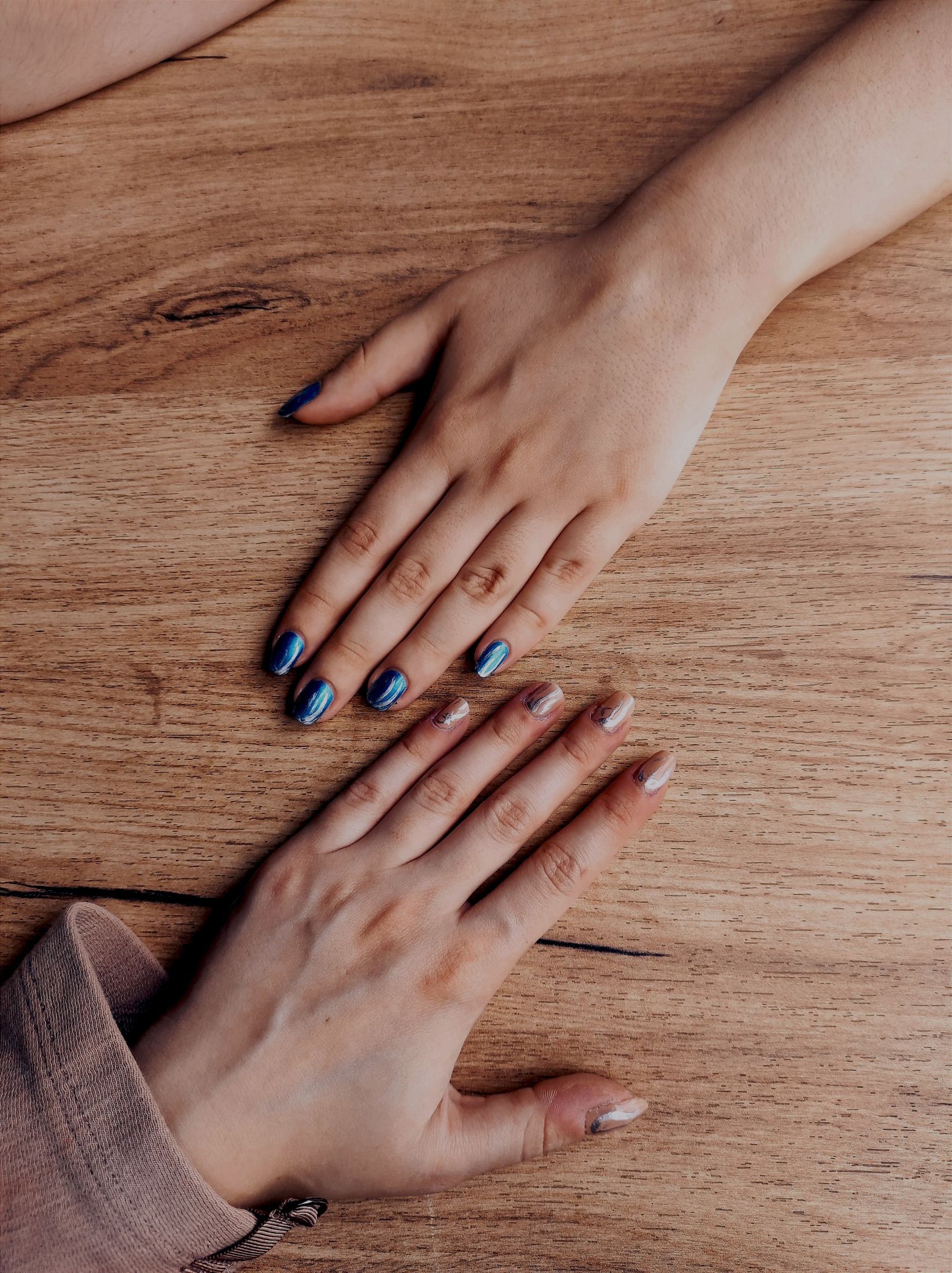 Two sets of hands with painted fingernails resting on a wooden tabletop, showing completed polish finishes and visible nail borders.