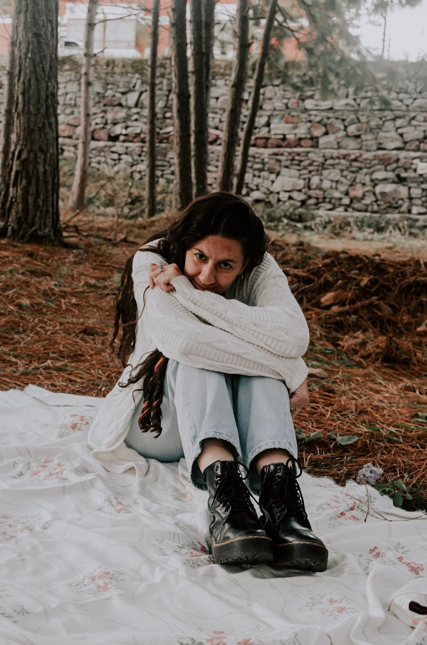 Woman seated on a blanket outdoors in a cream cable-knit sweater, pale straight-cut jeans ending above black lace-up boots; seated hem and boot opening are visible.