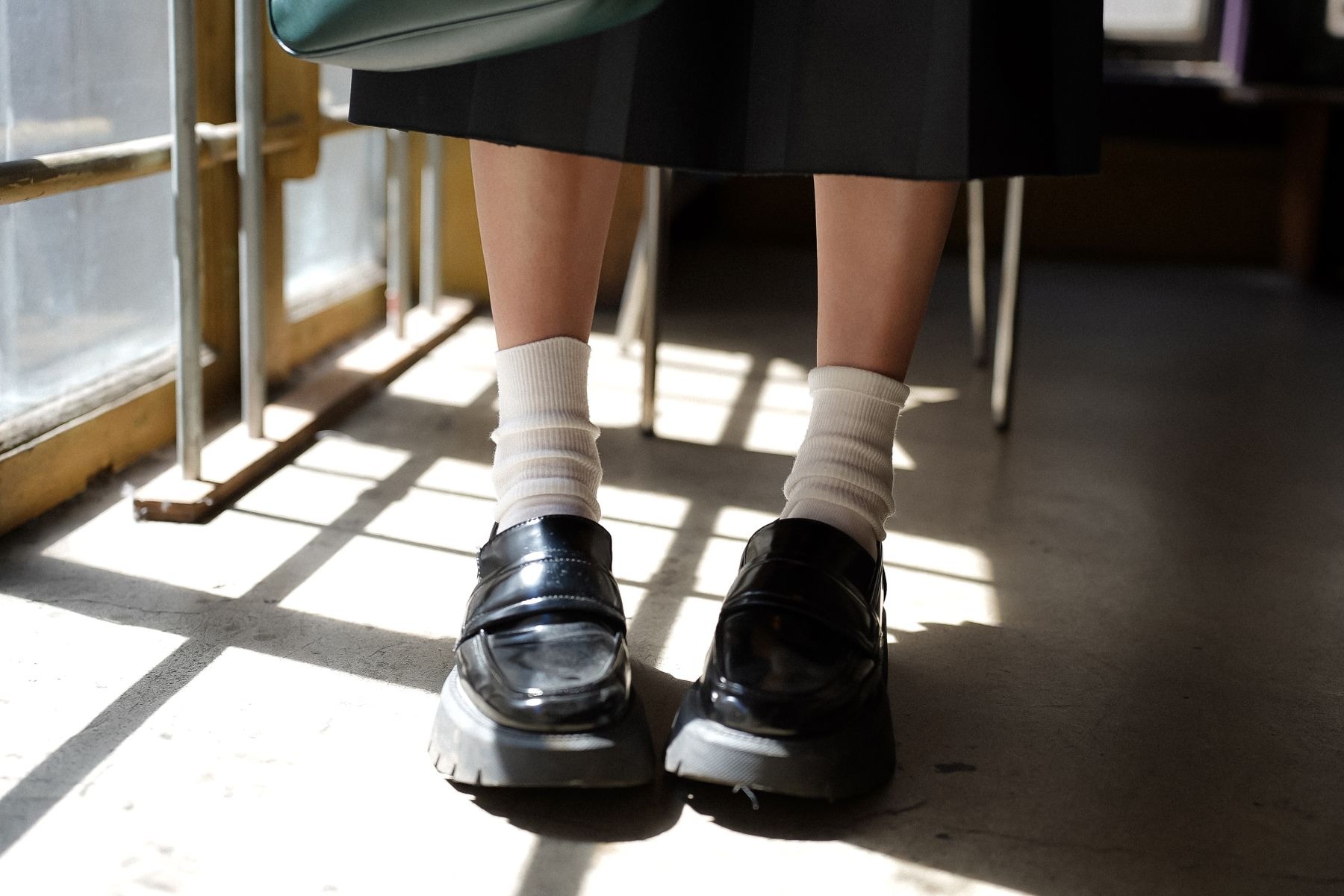 Black loafers worn with white socks beneath a dark skirt in window light, with a small green bag partly visible; sock-and-loafer comparison, not a trouser example.