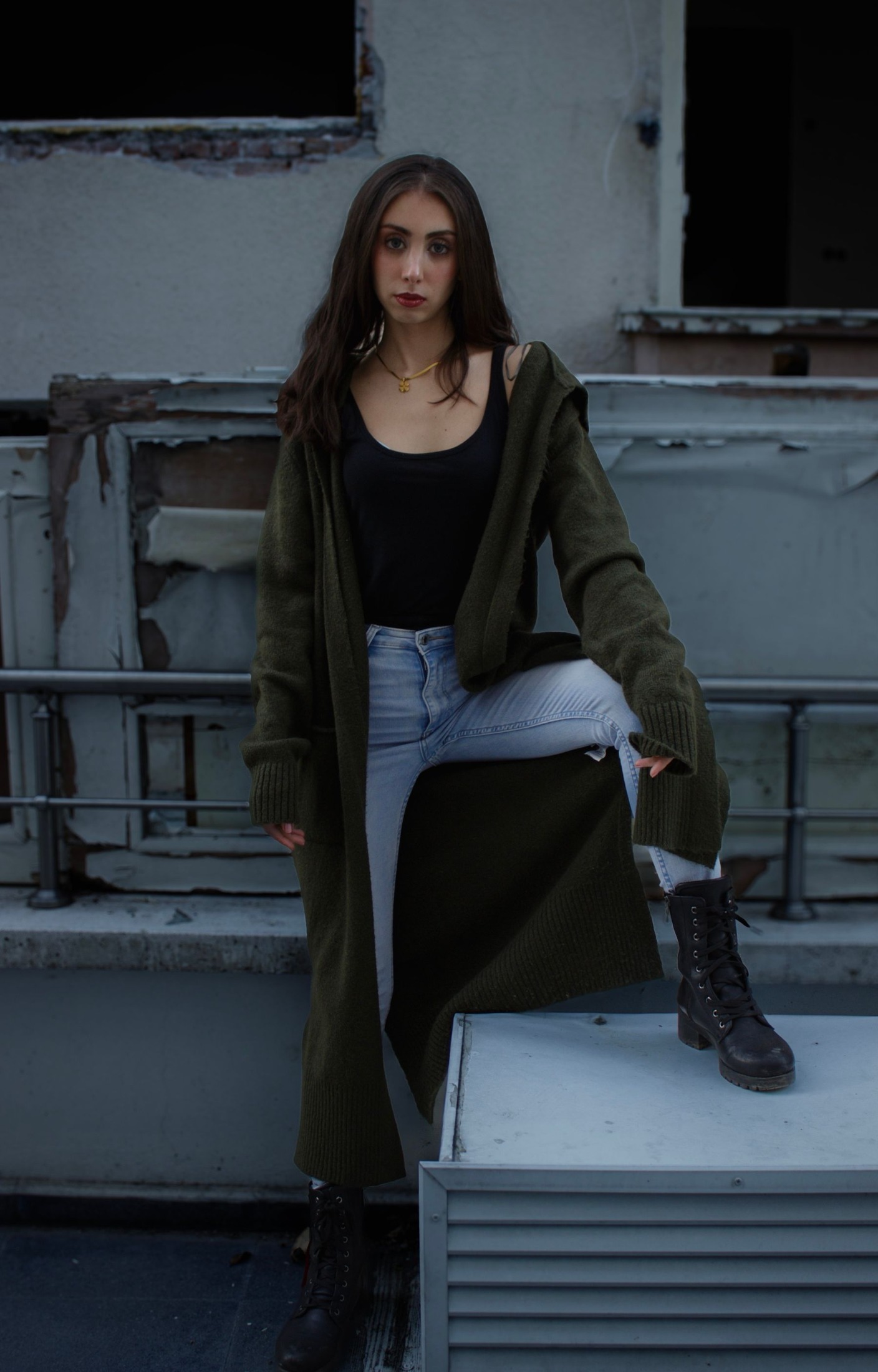 Woman wearing a long open-front olive cardigan over a black tank and pale ripped jeans, with black lace-up boots on gray steps.