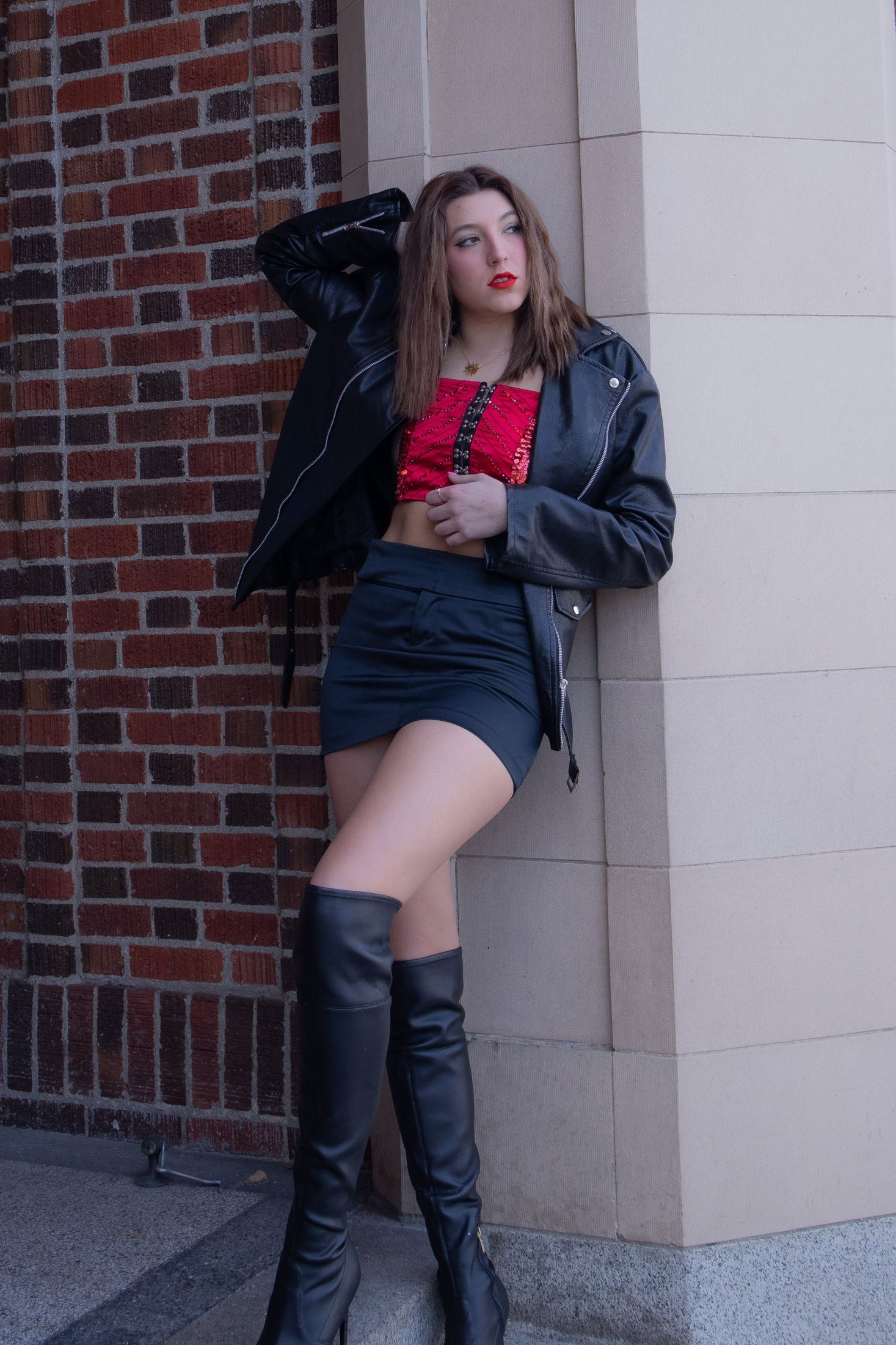Woman wearing black knee-high boots, a black mini skirt, red crop top and black leather jacket on a sidewalk.