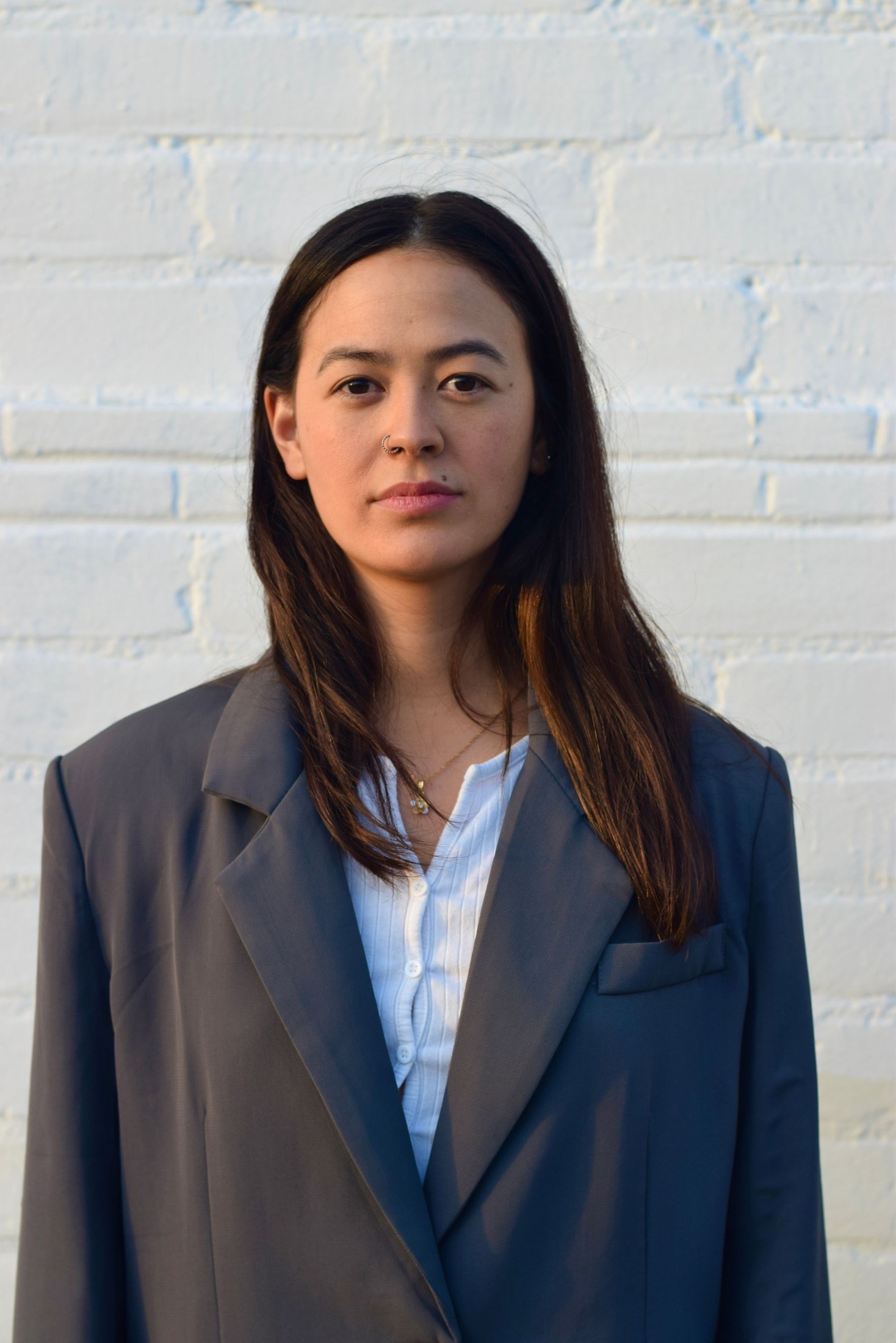 Woman wearing a loose gray blazer over a pale striped shirt against a white brick wall; upper-body portrait.