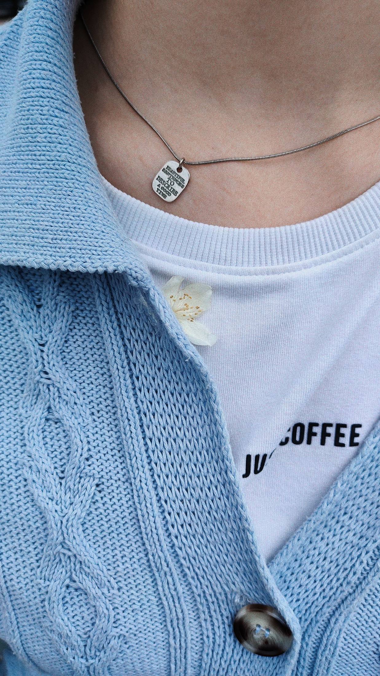 Close-up of an open blue cable-knit cardigan over a white printed crew-neck T-shirt, with a small pendant and white flower detail; neckline and layer construction visible.