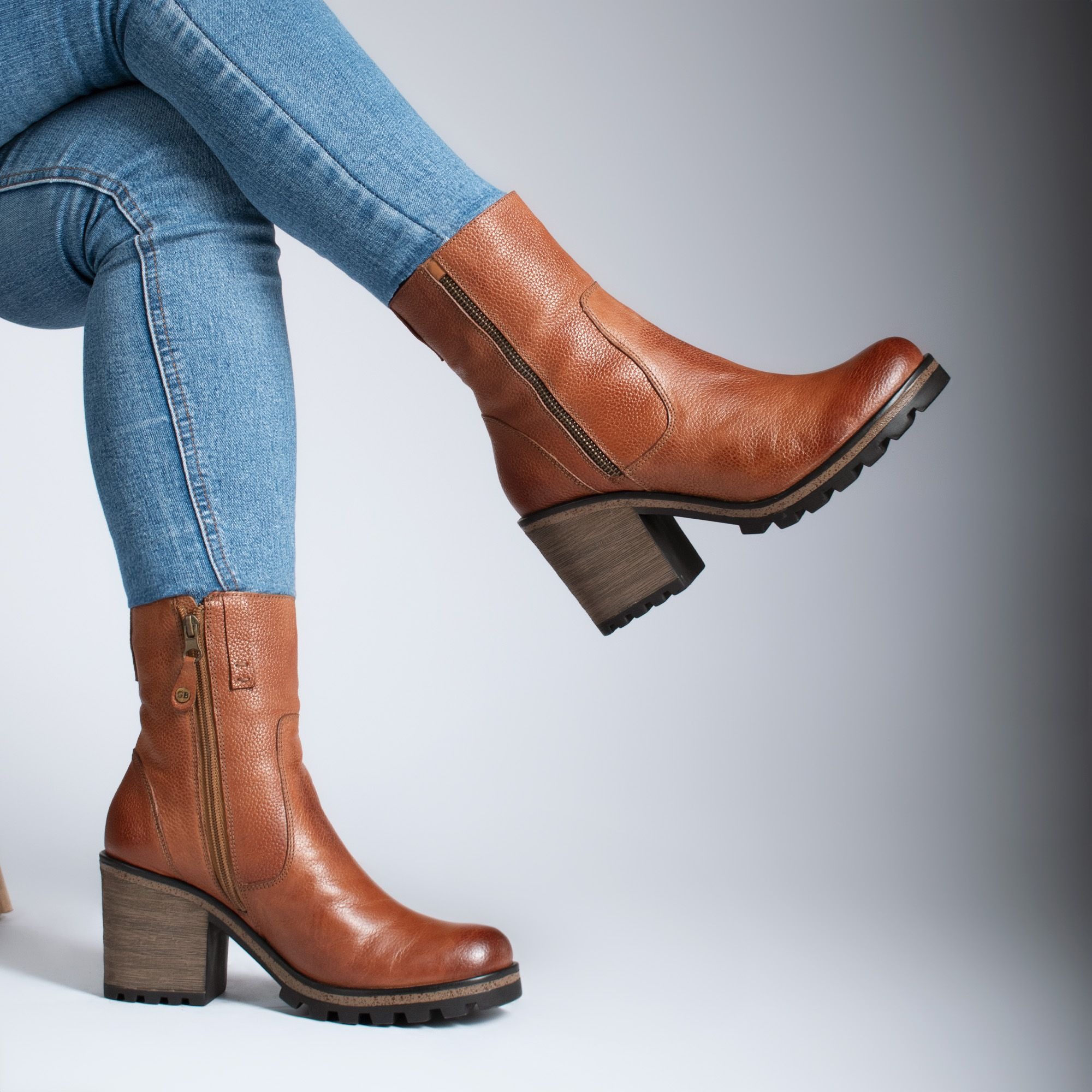 Close-up of brown side-zip ankle boots with block heels and visible tread, worn with fitted blue jeans against gray; footwear component illustration only, with no waterproof or grip-performance claim.