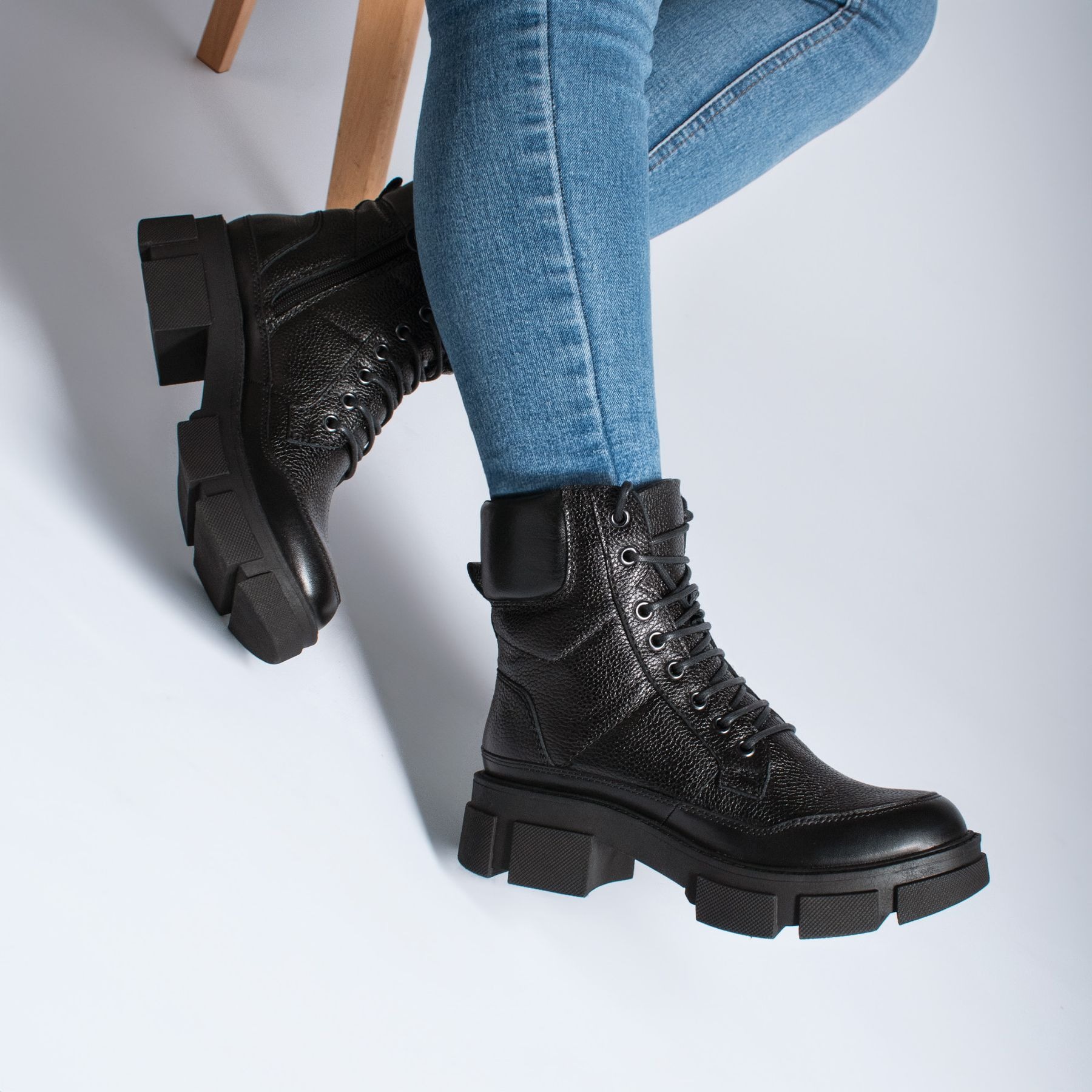 Black lace-up ankle boots with chunky lug soles worn with blue jeans against a pale studio floor.