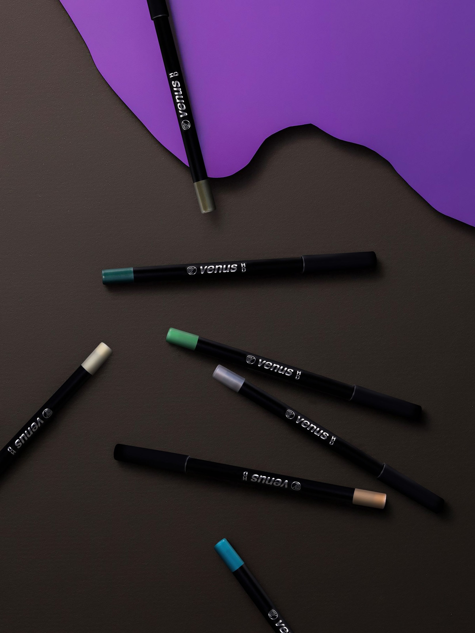 Several dark eyeliner pencils with differently colored end caps arranged diagonally on a dark brown surface with purple paper accents.