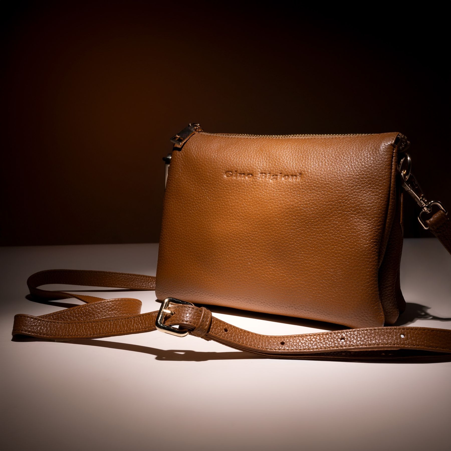 Brown rectangular crossbody bag on a tabletop with its long adjustable strap and buckle laid in front; strap hardware and attachment detail.