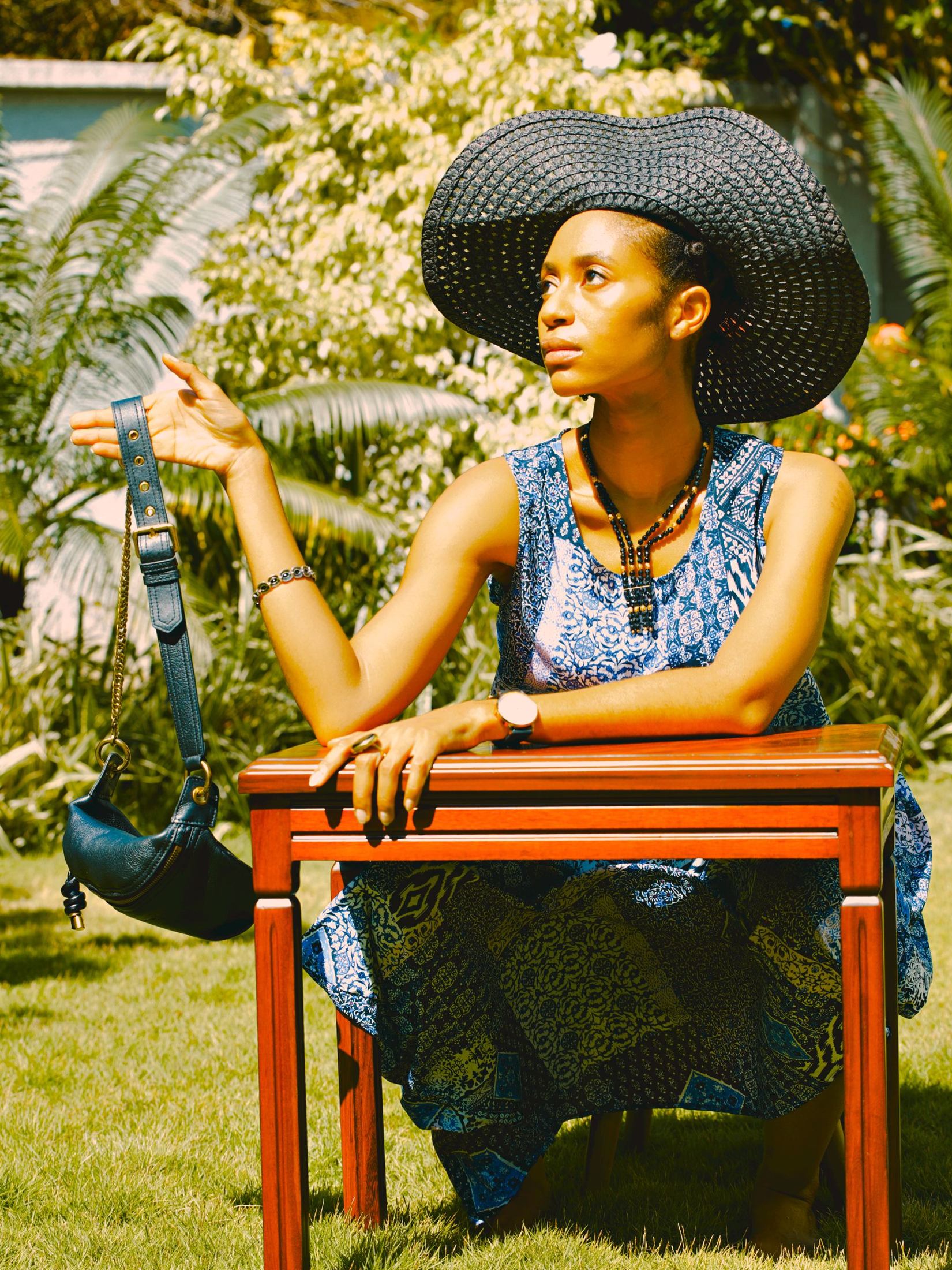Seated woman in a blue patterned sleeveless dress wearing a wide black woven brimmed hat in a garden, with a small black bag held beside her.