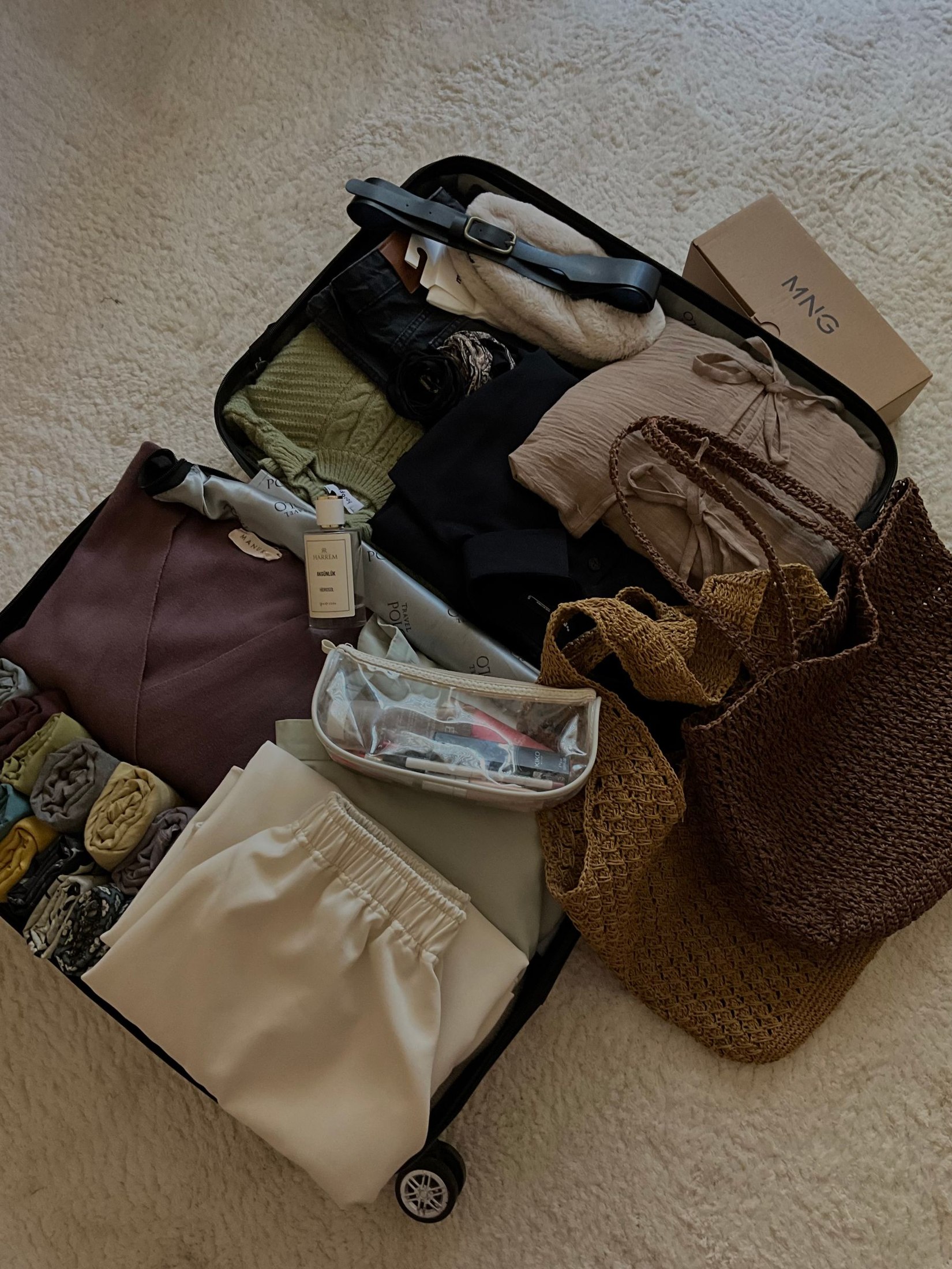 Overhead view of an open suitcase filled with folded pale and dark clothing on a gray textured floor, with small accessories and a woven bag also visible; packed-clothing example, not an exact capsule inventory.