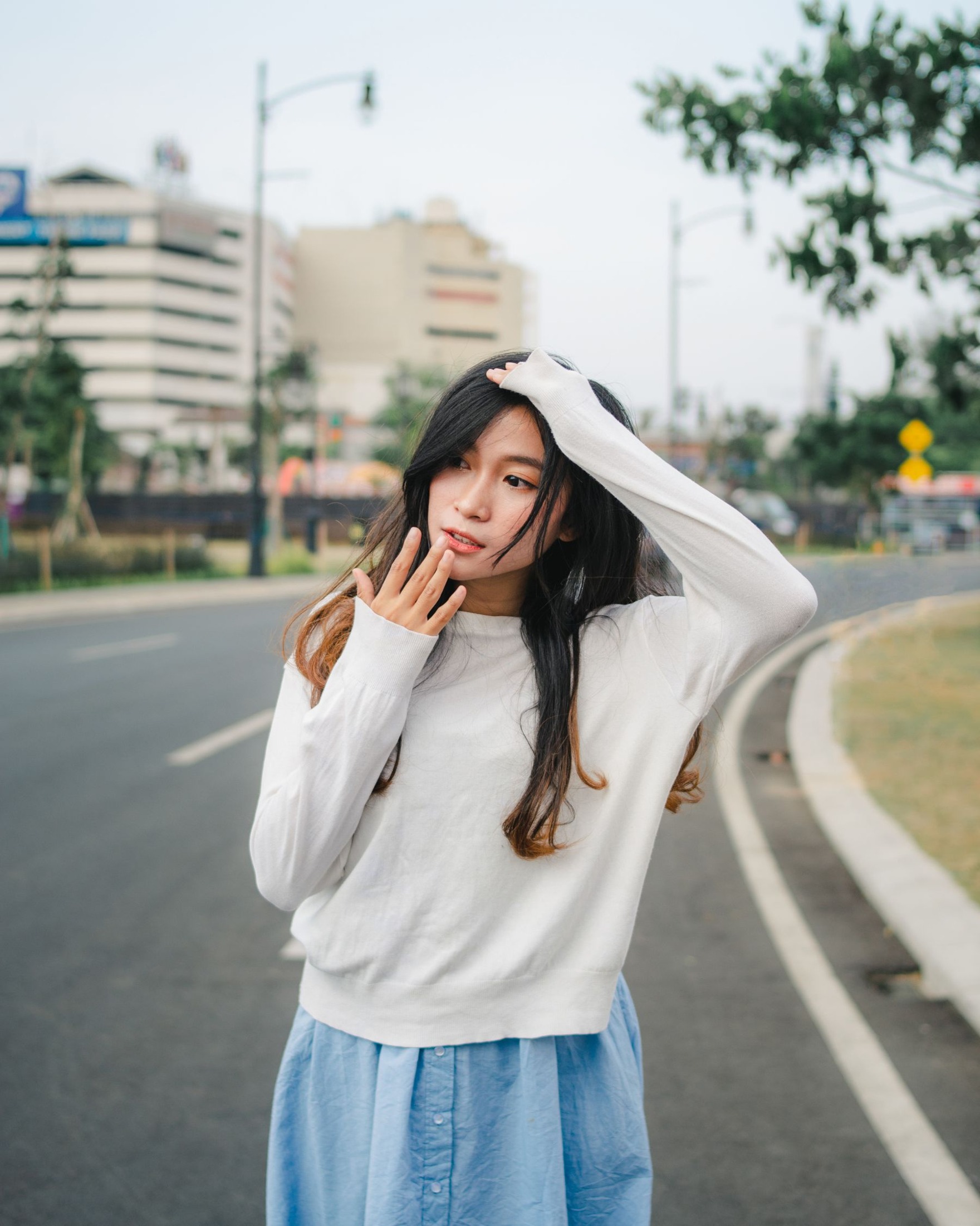 Woman wearing a plain white long-sleeved sweater with pale blue trousers beside a street.