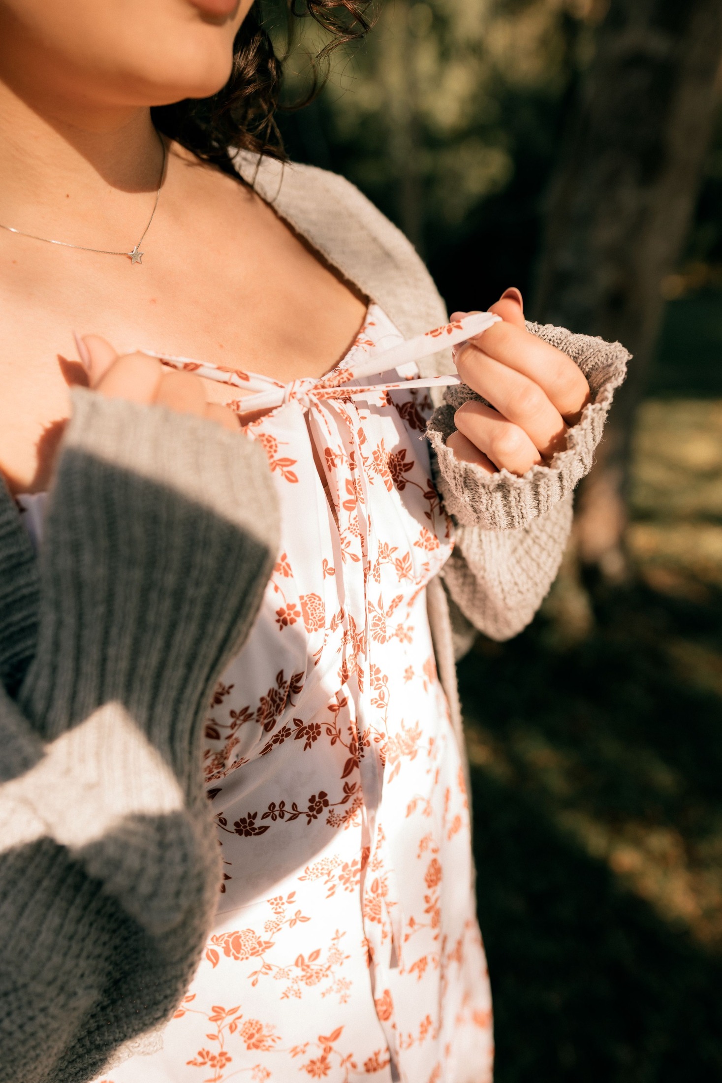 Close-up of a pale floral dress neckline and tie beneath an open gray-beige ribbed cardigan; the wearer's hands hold the dress tie, showing the meeting of the two layers.