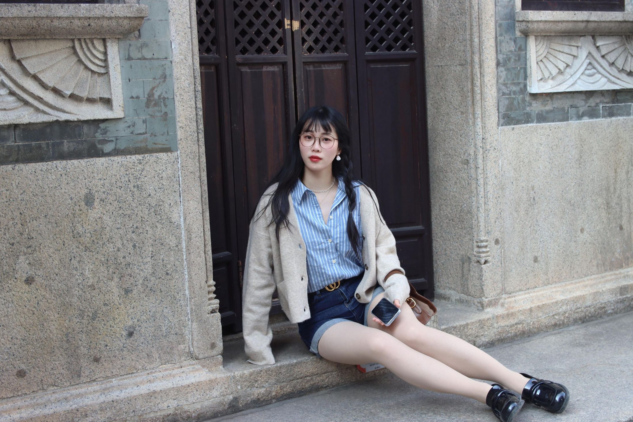 Woman seated in a cream cardigan over a blue-and-white striped shirt with dark navy denim shorts and black loafers; illustrates the navy-and-light base of a palette before adding green, which is not present in this outfit.