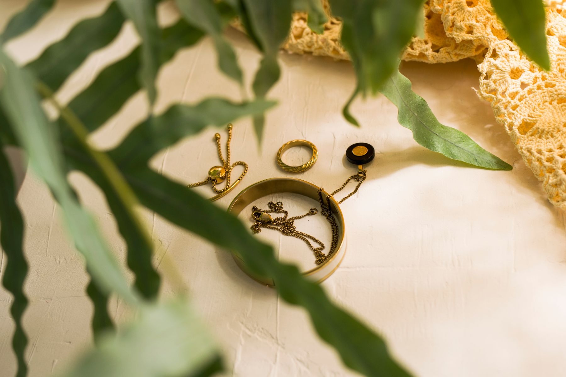 Gold-colored bangle, ring and fine-chain jewelry on a pale surface framed by green leaves and a crocheted textile.