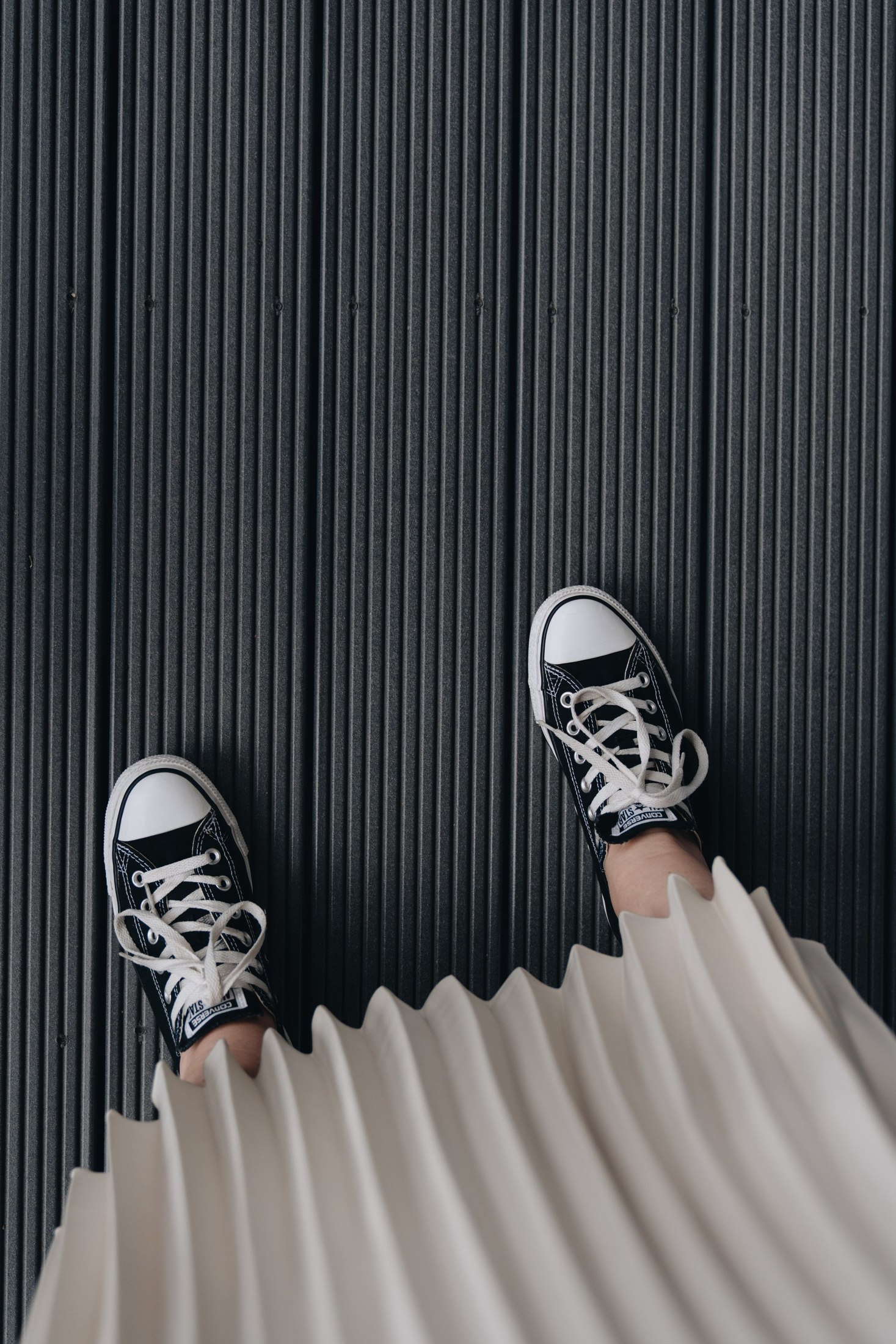Downward view of a plain light pleated skirt hem above black canvas lace-up trainers with white toe caps on a dark ribbed surface; skirt-and-shoe detail rather than a complete outfit.