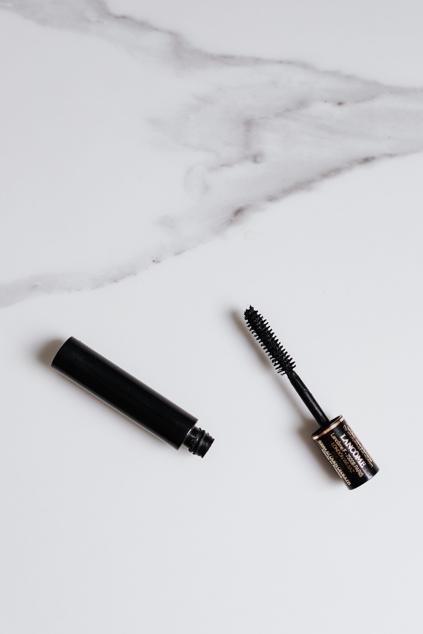 Open black mascara tube and its separate wand arranged on white-and-gray marble, with the brush clearly visible.