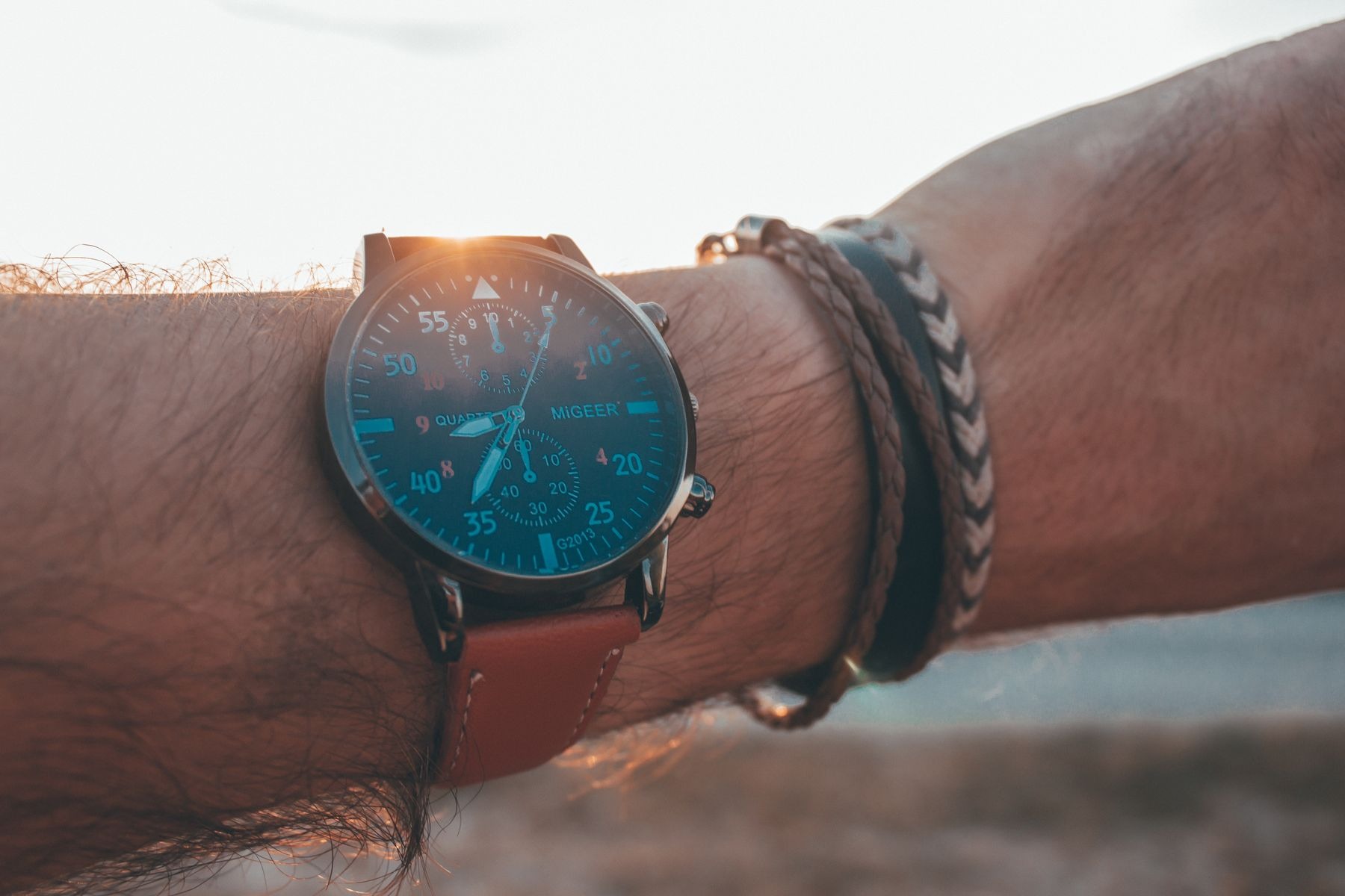 Close-up of a wrist wearing a blue-faced watch on a brown strap beside two bracelets, with low sunlight behind the wrist; contrasting watch and bracelet textures.