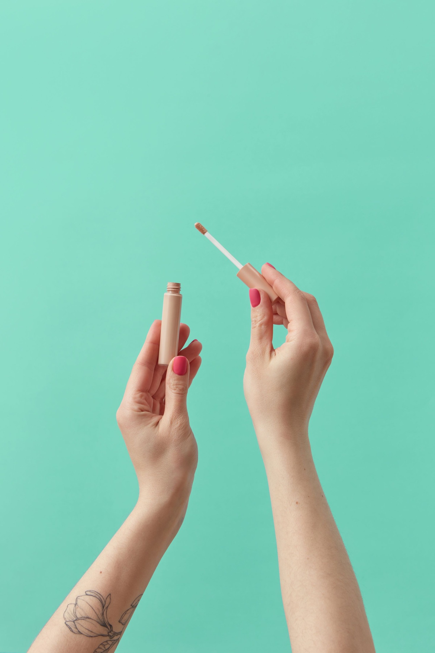 Hands holding an open beige concealer tube and its lightly coated doe-foot applicator against a mint-green background.