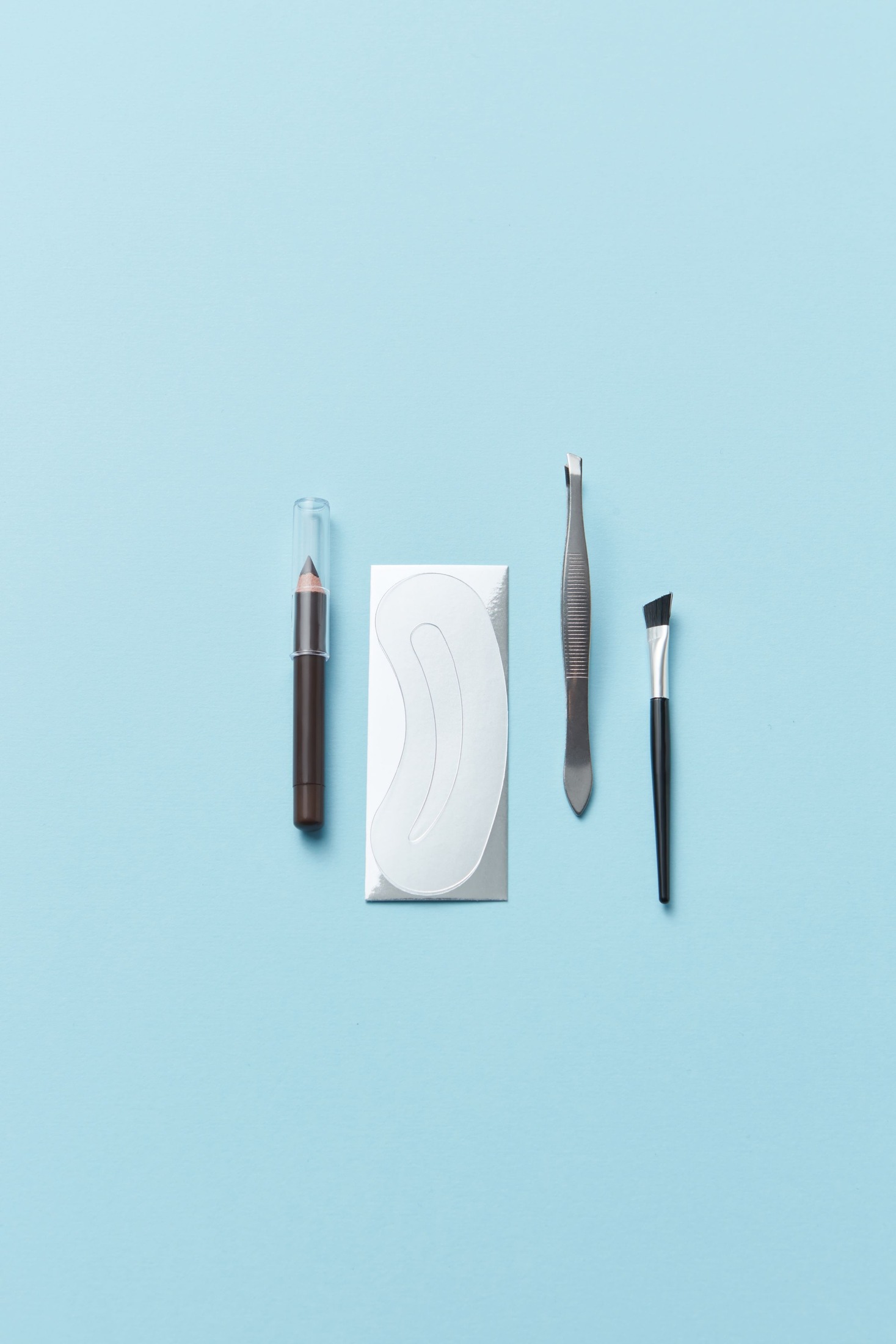 Capped sharpened brow pencil, eyebrow stencil card, tweezers, and an angled brow brush laid separately on a pale blue surface.