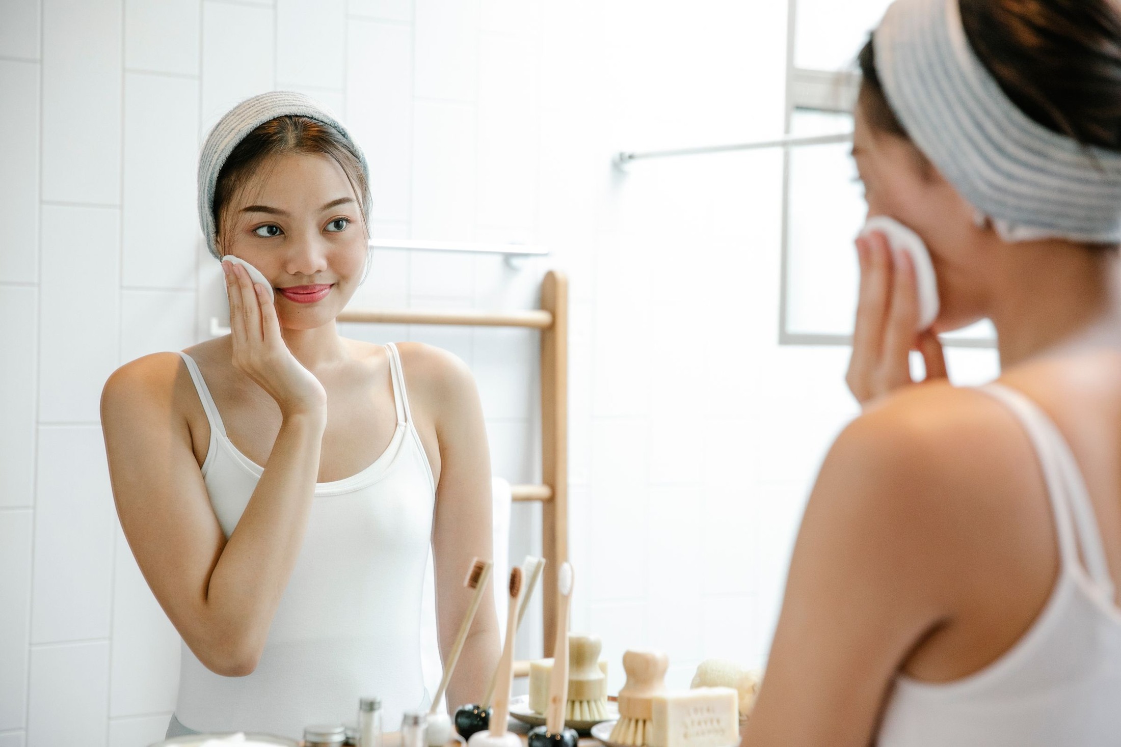 Woman using a white cotton pad on her face while looking at her reflection in a bathroom mirror.