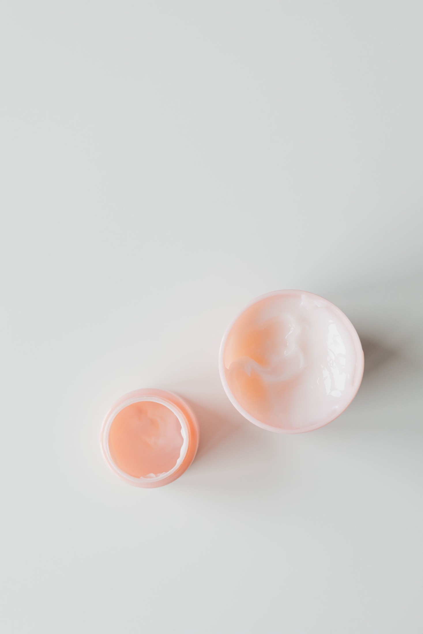 Overhead view of an open pale-pink cream jar and its lid with visibly swirled cream on a light gray surface; no serum bottles are shown.