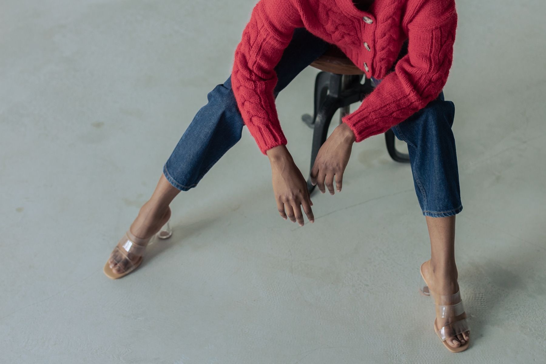 Cropped seated outfit showing a red cable-knit cardigan, blue jeans and clear-strap heeled sandals on a gray floor.