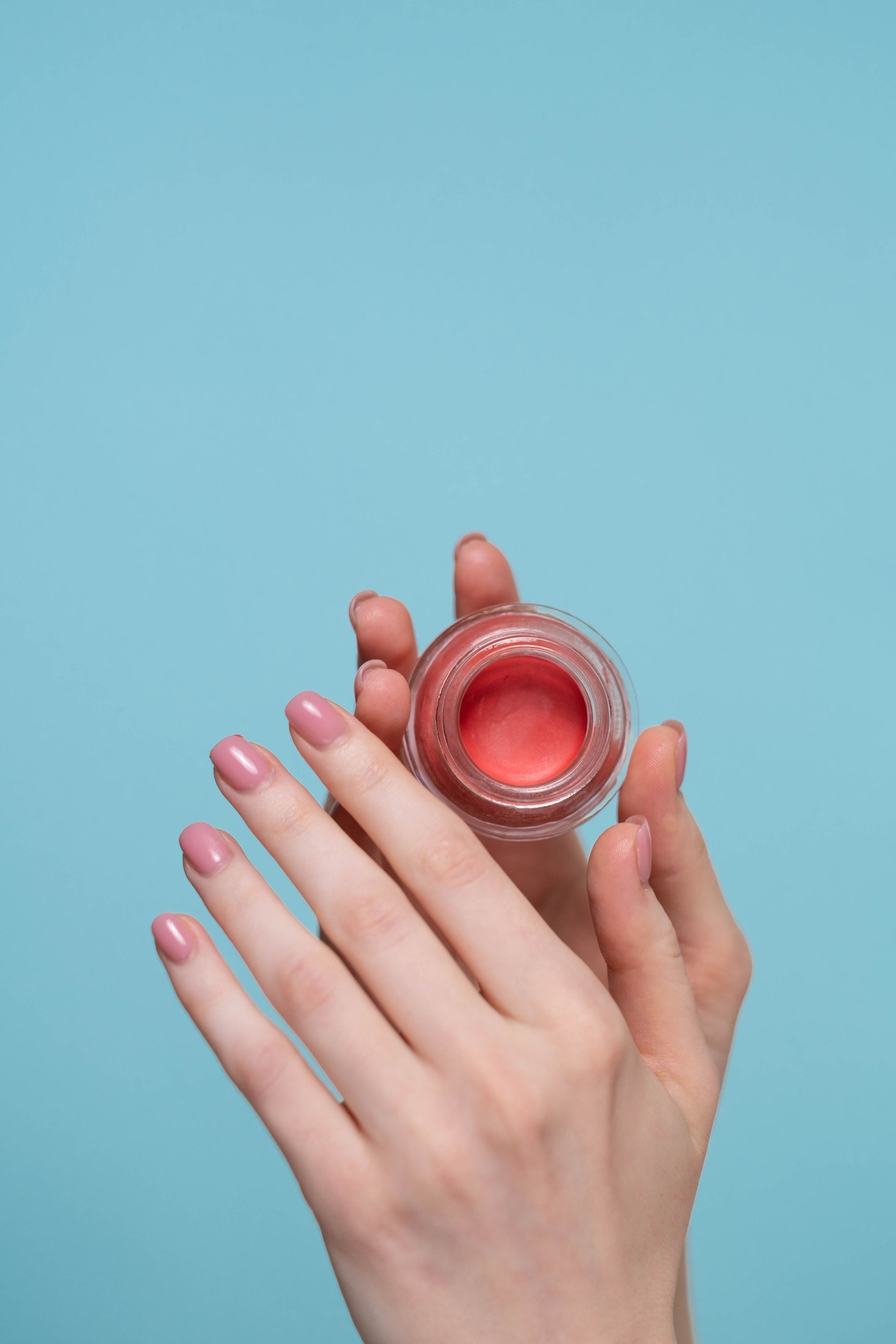 Hands holding an open small glass jar of coral-pink cream blush with its smooth creamy surface visible against a light blue backdrop.