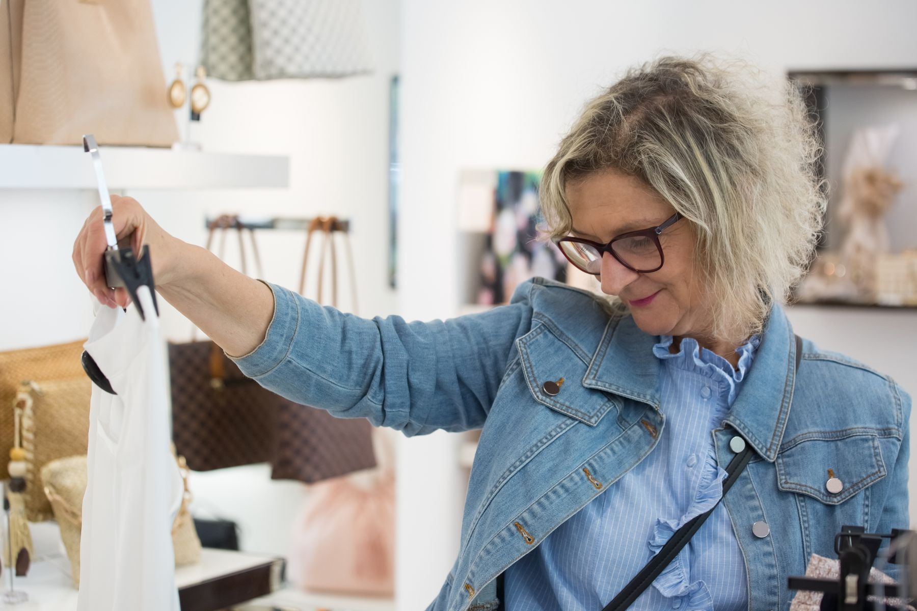 Older shopper in a denim jacket and glasses holding up a light garment on a hanger to examine it in a clothing shop; garment inspection illustration, shop's secondhand status not established.