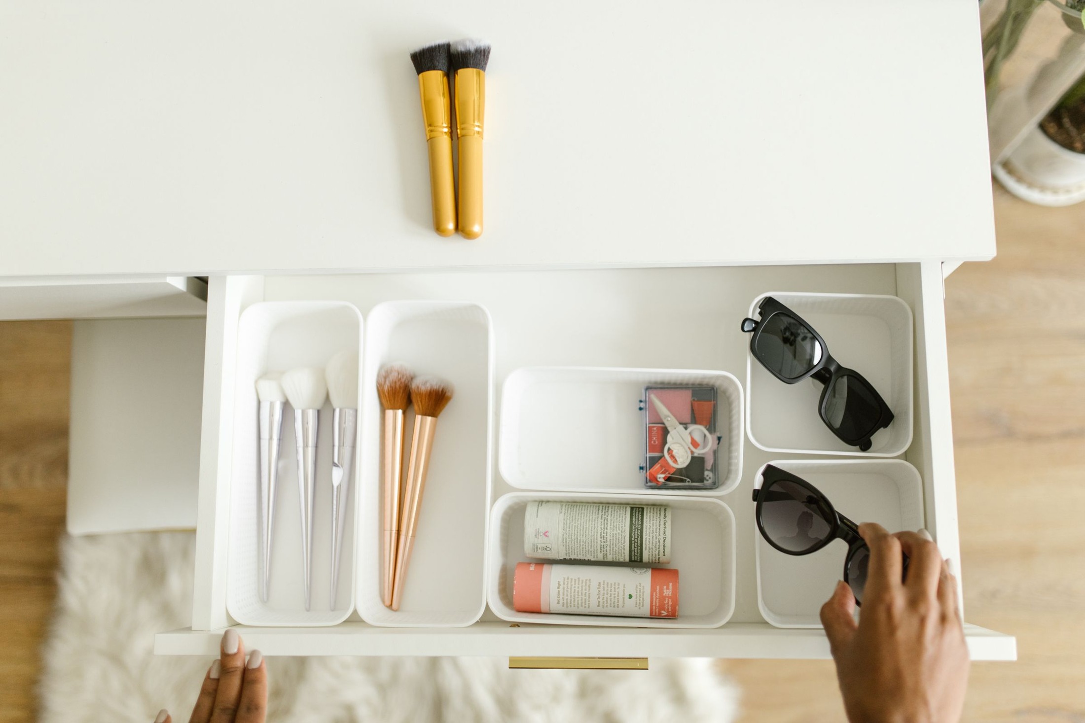 Open white drawer divided into shallow trays containing makeup brushes, small cosmetics, toiletry containers, and sunglasses, with a hand at the drawer edge.
