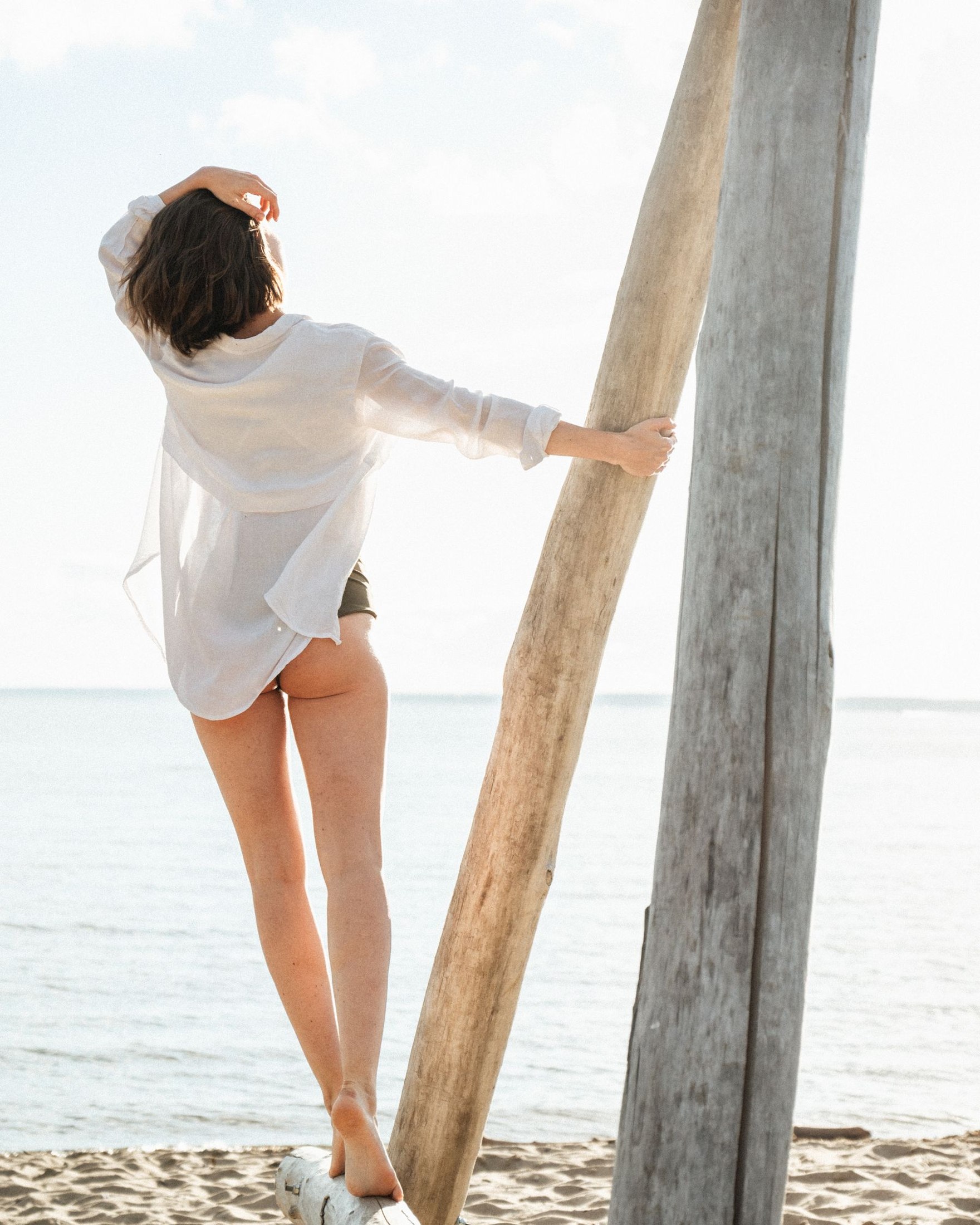 Rear view of a woman beside a tall wooden post wearing an open white long-sleeved shirt over a dark swimsuit at the beach; cover-up layer only.