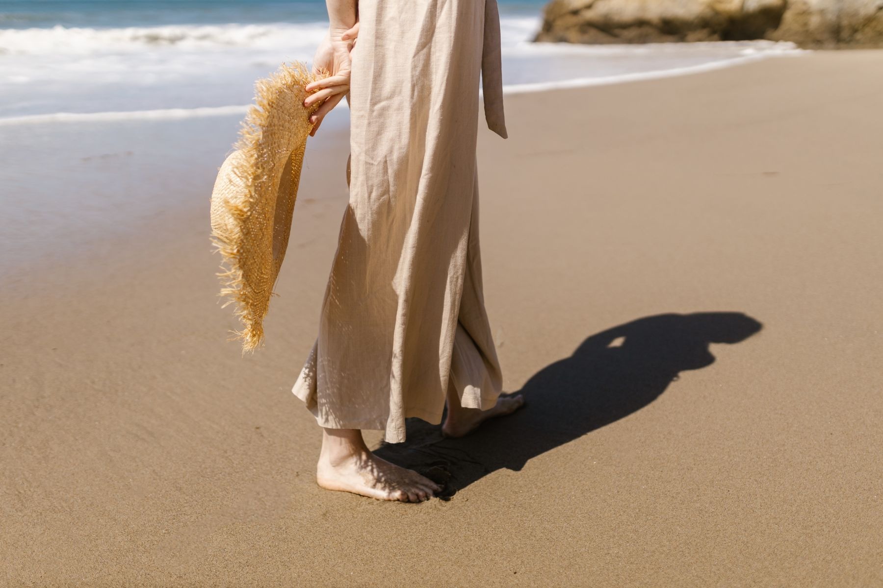 Close view of beige wide-leg linen pants worn barefoot on a beach, with a straw hat held at the side.