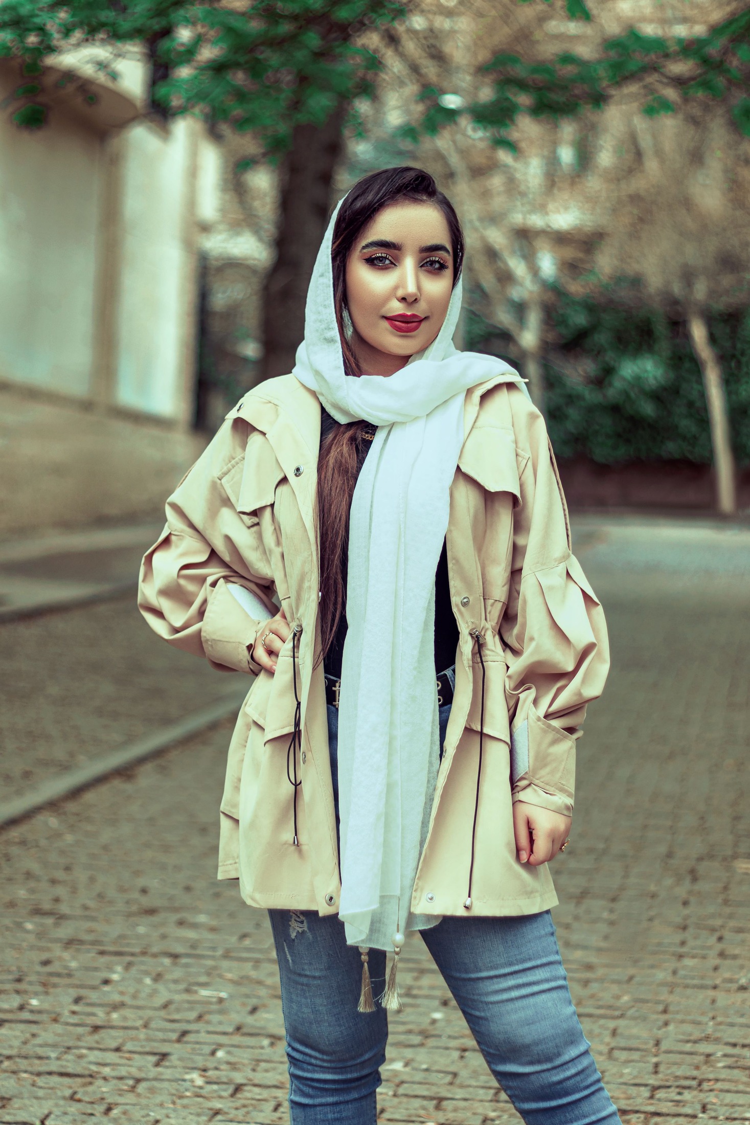 Woman wearing a light scarf over her hair and around the neck with long flat ends outside a beige drawstring coat; the open collar and scarf ends are visible.