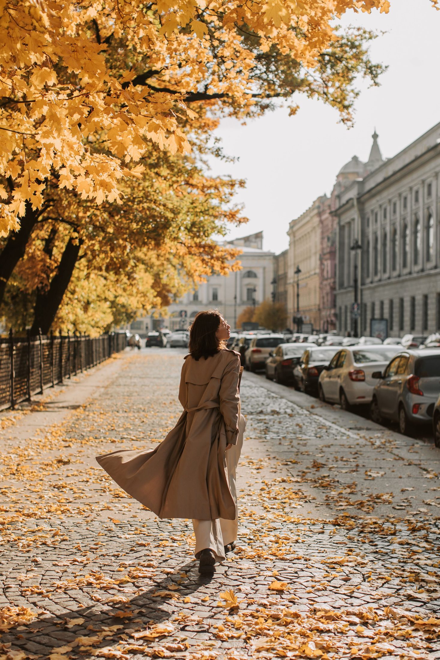 Rear full-length view of a woman walking on a leaf-covered sidewalk in a long beige trench with a belt tied behind, pale trousers and dark shoes.