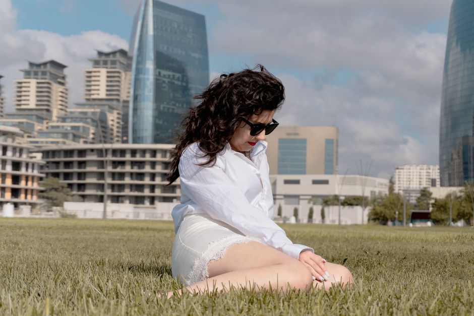 Woman in white attire sitting on grass in Baku with urban skyline backdrop.